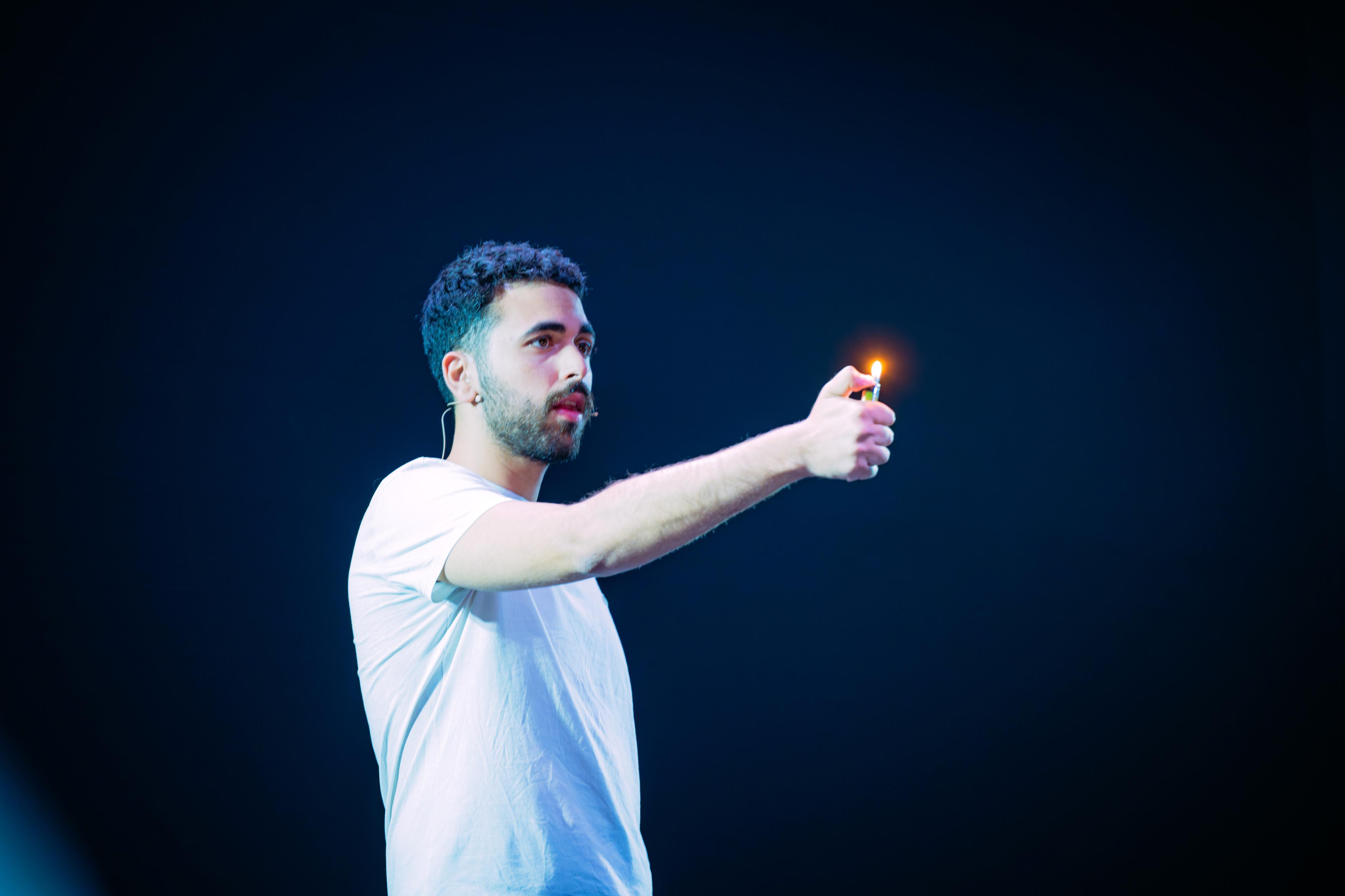 Fadi Murad on a dark stage, holding a lit lighter at arm’s length.