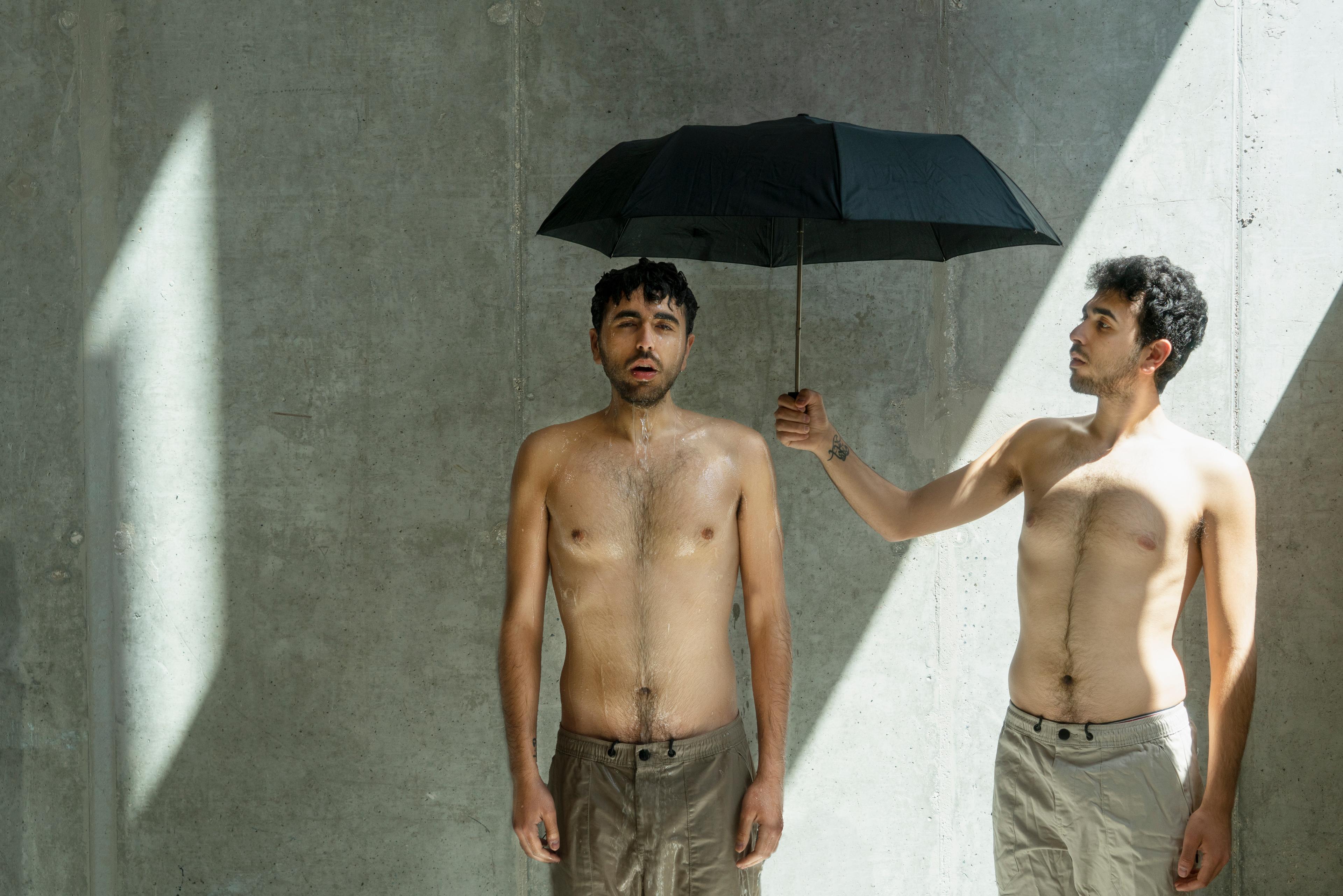 Come Back Home key art: against a sunlit concrete wall, Fadi Murad holds a black umbrella over a second, drenched Fadi Murad.