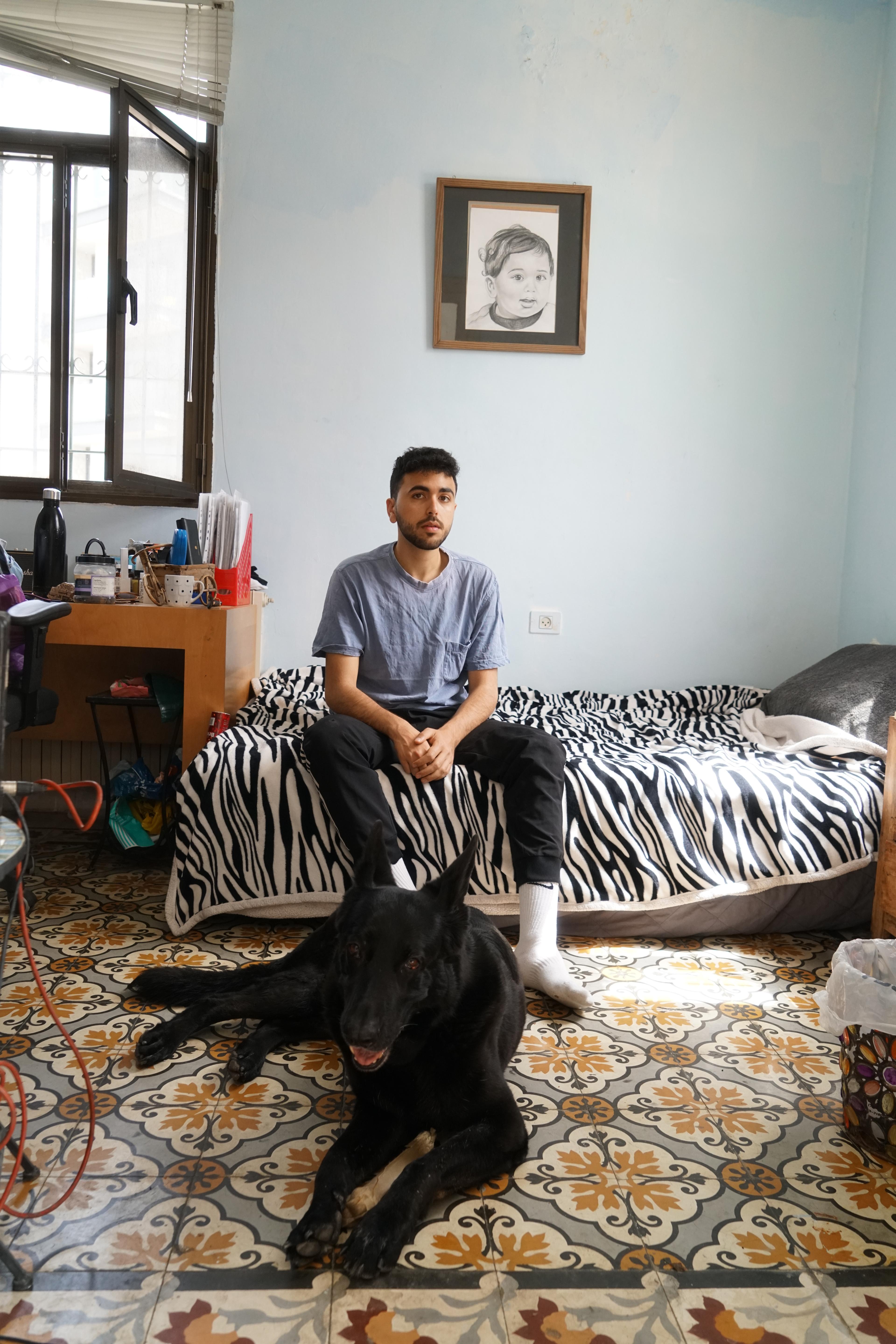 Fadi Murad sits on a zebra-print bed in a pale blue room, a black dog lying on the patterned tiles at his feet.